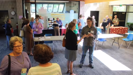 This is a photo of Presidents Hall at First UU of Richmond with several people standing in small groups, engaged in conversation and holding cups of coffee. Some people are standing at an information table near the back of the picture.