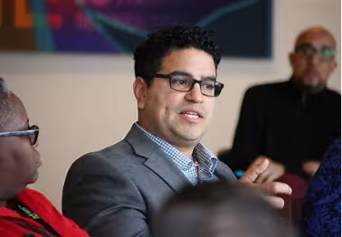 Dr. Ortega is a Hispanic with black curly hair black rimmed glasses. He is sitting down and wearing a blue button-down shirt opened at the collar. He is wearing a gray suit jacket and talking to other people in the picture. To his left there is an older black female with black glasses. To the right of Dr. Ortega there is an African-American man with a goatee and glasses in the foreground. Above Dr. Ortega there is a partial picture of a colored painting.