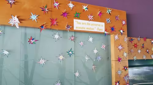 Wall decorated with colorful paper stars and a small sign with text. You are the universe in ecstatic motion. -Rumi