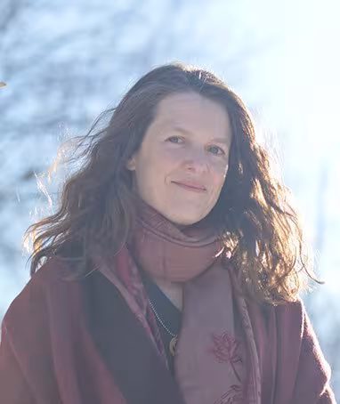 Person with long wavy hair, wearing a scarf and coat, standing outdoors in sunlight.