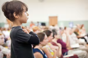 A young child with dark hair stands with arms crossed, facing left, in front of an out-of-focus seated audience in a public gathering.